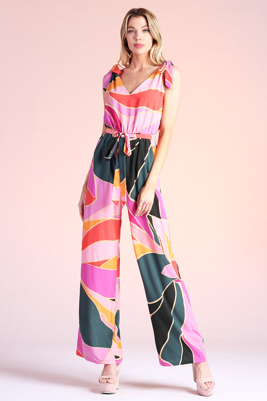 Blonde model leaning to the side, wearing pink, green and orange metamorphosis printed jumpsuit complete with ties on shoulders and pockets
