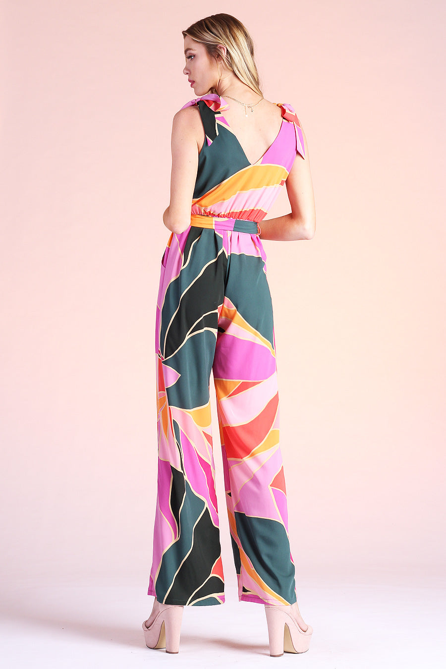 Blonde model posing backwards, wearing pink, green and orange metamorphosis printed jumpsuit complete with ties on shoulders and pockets