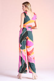 Blonde model posing backwards, wearing pink, green and orange metamorphosis printed jumpsuit complete with ties on shoulders and pockets