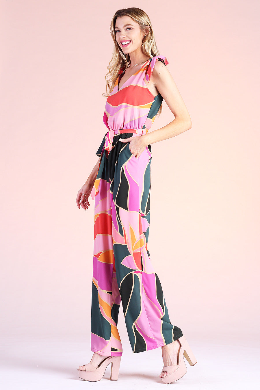 Blonde model posing wearing pink, green and orange metamorphosis printed jumpsuit complete with ties on shoulders and pockets