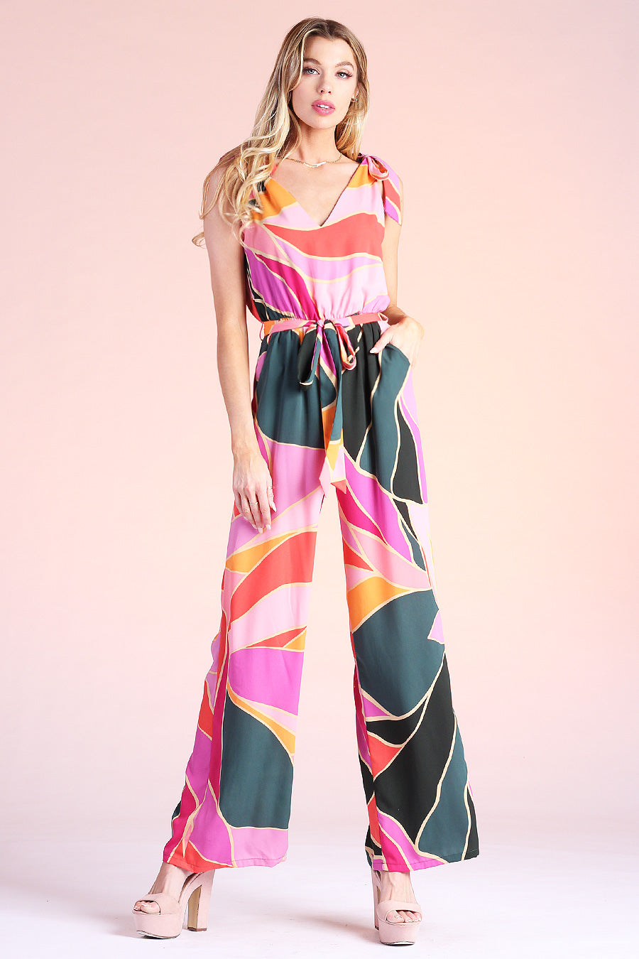 Blonde model posing wearing pink, green and orange metamorphosis printed jumpsuit, complete with ties on shoulders and pockets